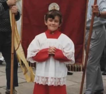 Altar boy in red cassock standing in front of a banner at a Blessed Sacrament procession